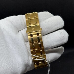 May include: A gold-toned watch band with a clasp, featuring a repeating rectangular link design. The band is held by a gloved hand against a dark background. A white string is attached to the band.