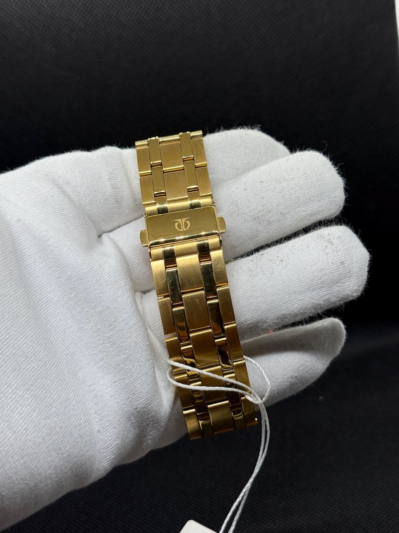 May include: A gold-toned watch band with a clasp, featuring a repeating rectangular link design. The band is held by a gloved hand against a dark background. A white string is attached to the band.