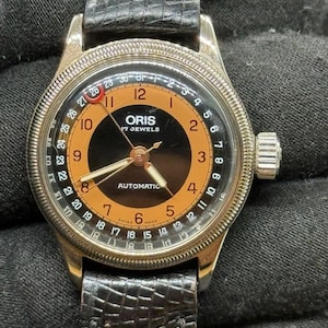 Puede incluir: Un reloj de pulsera automático Oris con esfera negra y naranja, que presenta una aguja de fecha con punta de media luna roja, números arábigos y una correa de cuero negro texturizado. La esfera muestra el texto Oris, 17 Jewels y Automatic.