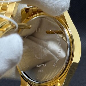 May include: Close-up of a gold-toned wristwatch. The watch has a gold band and a circular face. The back of the watch is visible, with the text "STAINLESS STEEL BACK" and "1627YAA INDIA".