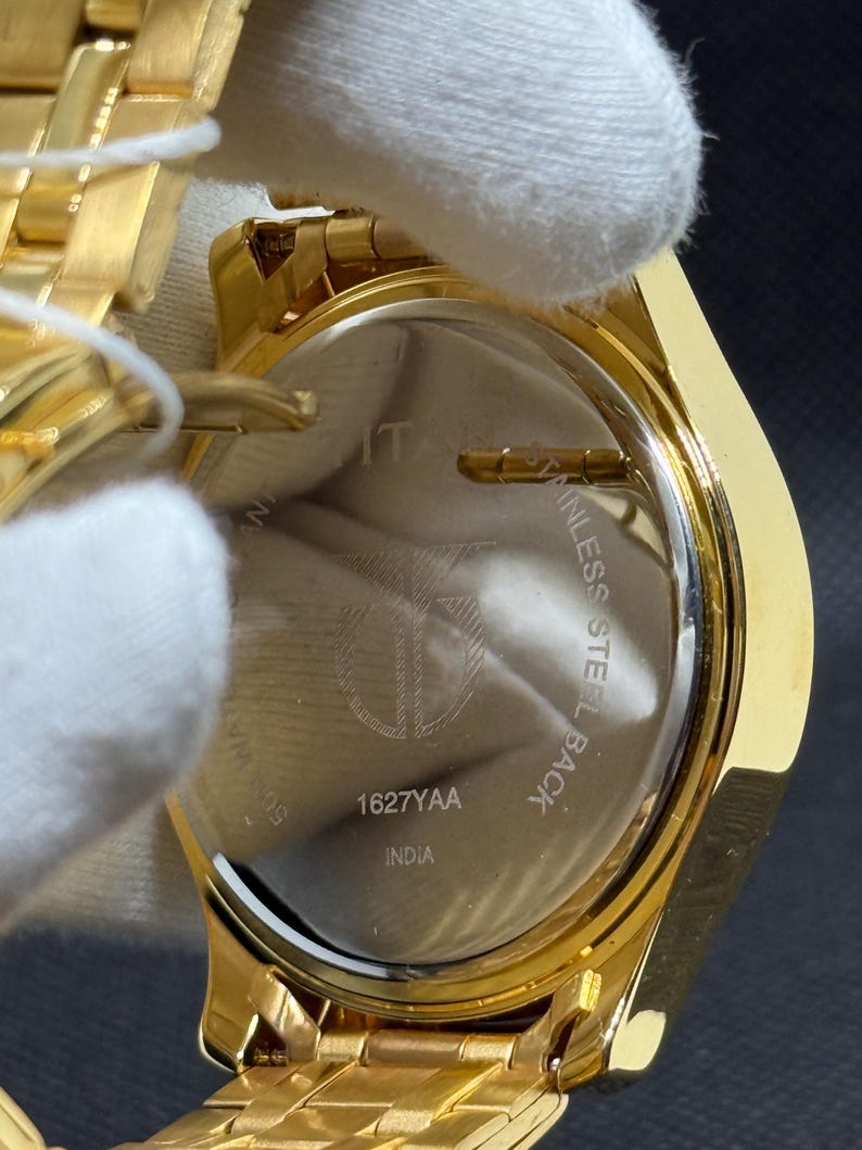 May include: Close-up of a gold-toned wristwatch. The watch has a gold band and a circular face. The back of the watch is visible, with the text "STAINLESS STEEL BACK" and "1627YAA INDIA".
