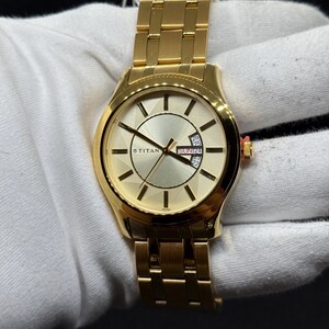 May include: A gold-toned wristwatch with a matching band. The watch face is a light gold color with black hands and markers. The watch has a date display and the brand name "TITAN" is visible. The watch is on a white surface.