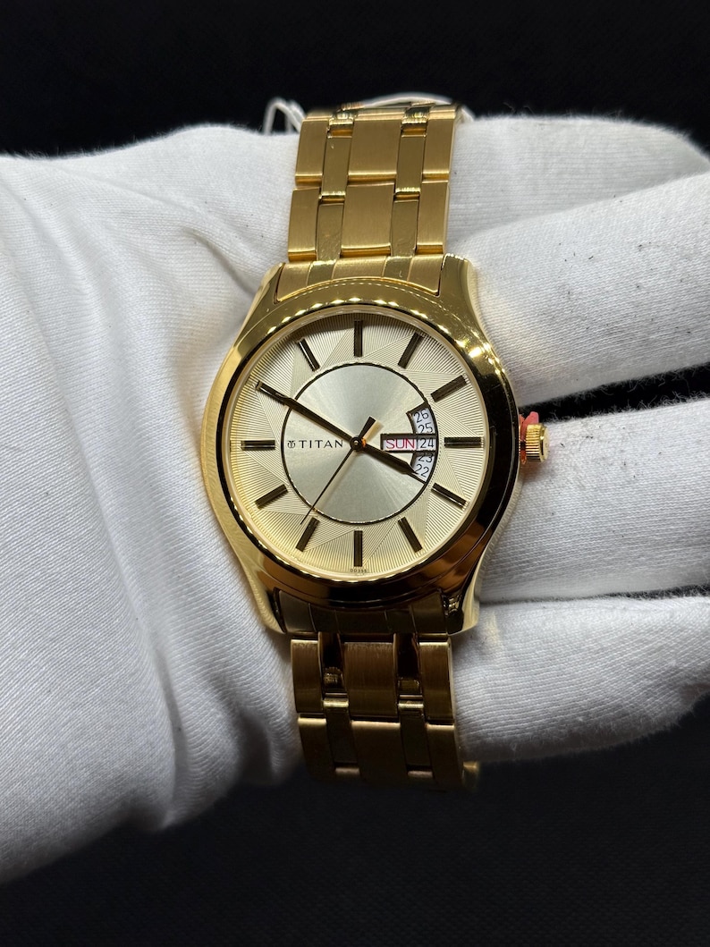May include: A gold-toned wristwatch with a matching band. The watch face is a light gold color with black hands and markers. The watch has a date display and the brand name "TITAN" is visible. The watch is on a white surface.
