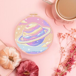 May include: A pastel-colored cross-stitch embroidery of a planet with rings, set in a round wooden hoop. The planet is in shades of blue, purple, yellow, and pink. The background includes clouds and stars. The image is on a pink surface.