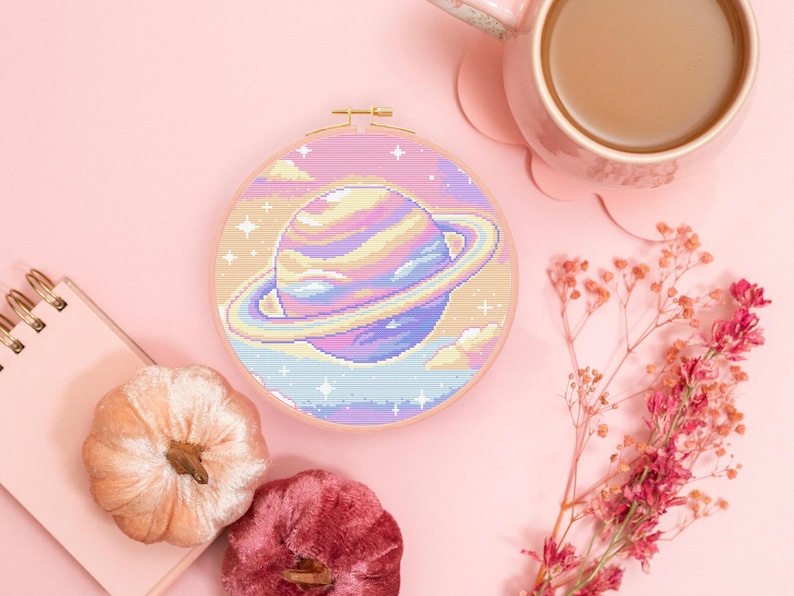 May include: A pastel-colored cross-stitch embroidery of a planet with rings, set in a round wooden hoop. The planet is in shades of blue, purple, yellow, and pink. The background includes clouds and stars. The image is on a pink surface.
