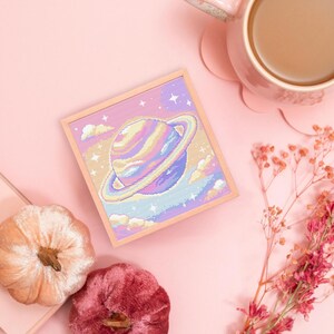May include: A pastel-colored cross-stitch artwork of a planet with a ring, set against a starry sky and clouds. The artwork is in a light wooden frame. The image also includes a pink mug with coffee, two velvet pumpkins, a notepad, and dried flowers.