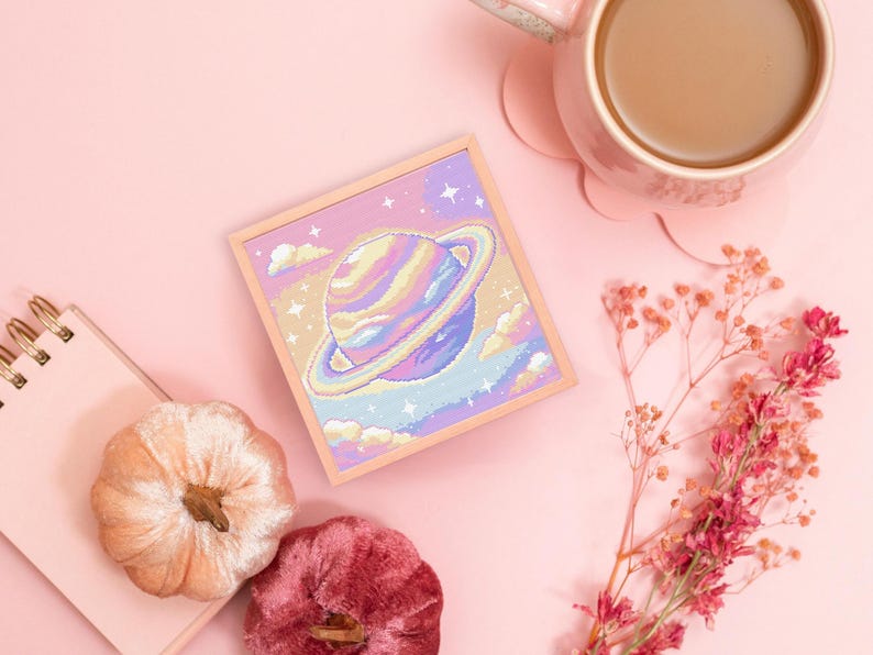 May include: A pastel-colored cross-stitch artwork of a planet with a ring, set against a starry sky and clouds. The artwork is in a light wooden frame. The image also includes a pink mug with coffee, two velvet pumpkins, a notepad, and dried flowers.
