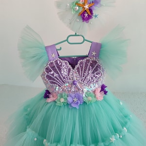Puede incluir: Un vestido de tul turquesa con un corpiño de concha púrpura, adornado con lentejuelas y perlas. El vestido tiene mangas de tul y una diadema a juego con una estrella de mar y perlas.
