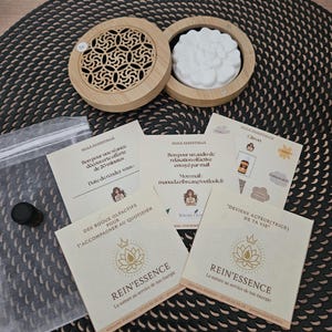 Peut inclure: Un diffuseur d'aromathérapie en bois avec un motif floral, ainsi qu'un insert blanc en forme de fleur. Plusieurs cartes avec du texte, dont "REIN'ESSENCE" et "HUILE ESSENTIELLE", sont éparpillées. Une petite bouteille noire et un sac en plastique transparent sont également présents.