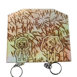 Può includere: Set di portachiavi in legno con Joker e Harley Quinn. Lo sfondo è inciso con "HAHA" ripetuto. I portachiavi sono attaccati a una base di legno con un anello di metallo.