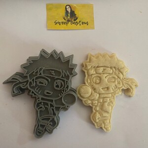 Può includere: Un tagliabiscotti in plastica grigia e un biscotto cotto a forma di personaggio dei cartoni animati con capelli a punta e una fascia. Il tagliabiscotti è accanto al biscotto cotto. Un piccolo adesivo giallo con la scritta "sweep custom" è sullo sfondo.