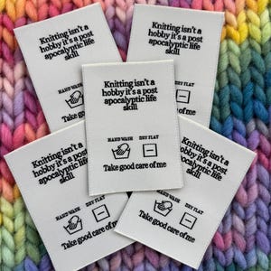 Puede incluir: Cinco etiquetas de tela blanca con texto negro: "Tejer no es un hobby, es una habilidad de vida post-apocalíptica." Instrucciones de cuidado: lavar a mano y secar en plano.