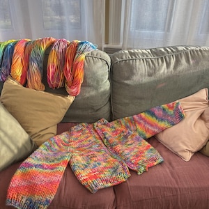 May include: A colorful, hand-knitted sweater in progress, featuring a vibrant rainbow of colors. The sweater is laid out on a mauve surface, with skeins of matching yarn draped over the back of a gray sofa. The scene is illuminated by natural light.