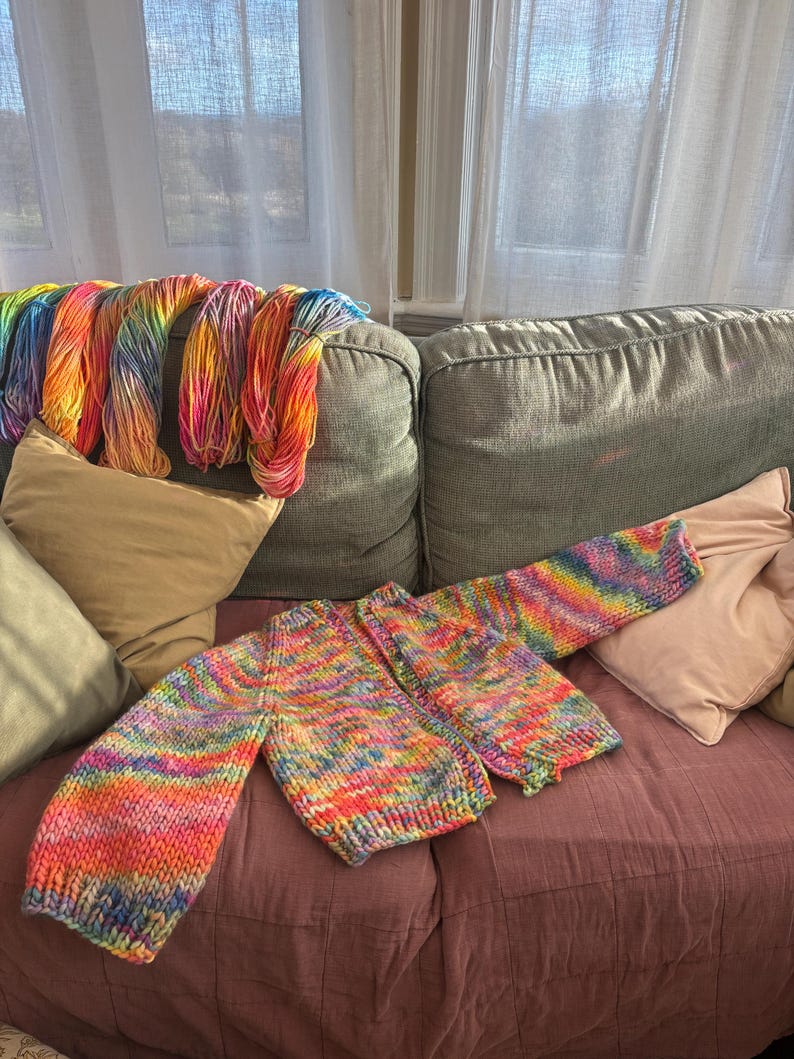 May include: A colorful, hand-knitted sweater in progress, featuring a vibrant rainbow of colors. The sweater is laid out on a mauve surface, with skeins of matching yarn draped over the back of a gray sofa. The scene is illuminated by natural light.