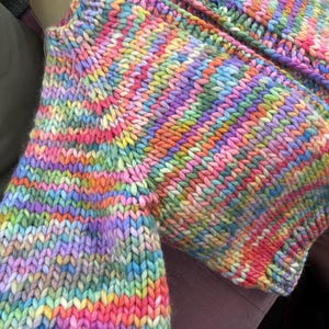 May include: A close-up of a hand-knitted garment in a vibrant rainbow of colors. The chunky knit features stripes of pink, orange, yellow, green, blue, and purple. The texture of the yarn is clearly visible.