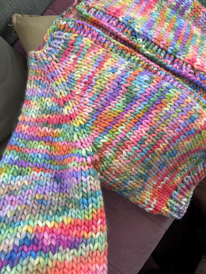 May include: A close-up of a hand-knitted garment in a vibrant rainbow of colors. The chunky knit features stripes of pink, orange, yellow, green, blue, and purple. The texture of the yarn is clearly visible.