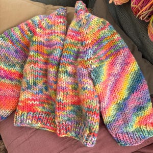 May include: A chunky knit cardigan in a vibrant rainbow of colors, including pink, yellow, blue, and purple. The open-front sweater is displayed on a mauve pillow, showcasing its bold, striped pattern and cozy texture. The sleeves are long and voluminous.