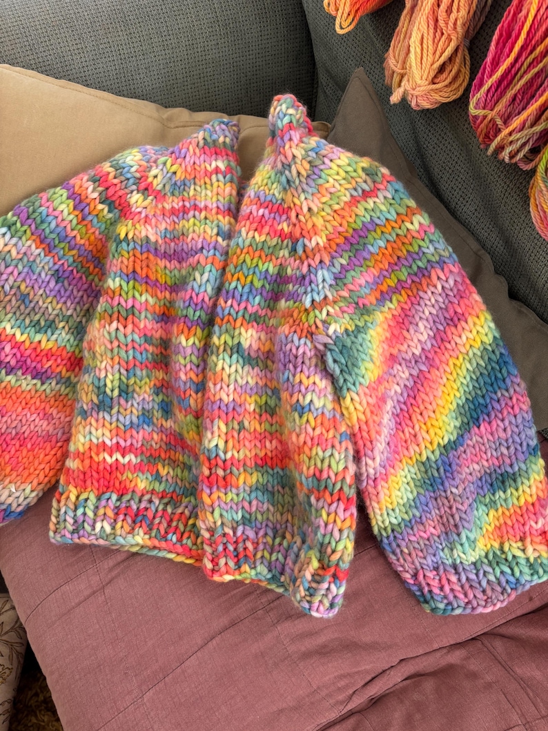 May include: A chunky knit cardigan in a vibrant rainbow of colors, including pink, yellow, blue, and purple. The open-front sweater is displayed on a mauve pillow, showcasing its bold, striped pattern and cozy texture. The sleeves are long and voluminous.