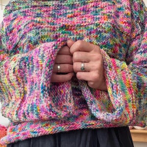 Könnte beinhalten: Ein handgestrickter Pullover mit langen Ärmeln und einem lebendigen, mehrfarbigen Garnmuster in den dominierenden Farben Pink, Blau, Gelb und Grün. Das Kleidungsstück hat einen weiten U-Boot-Ausschnitt und lockere Ärmel mit gerollten Bündchen.