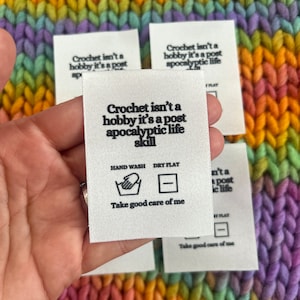Puede incluir: Etiqueta de tela blanca con texto negro e iconos. El texto dice "Crochet isn't a hobby it's a post apocalyptic life skill". Los iconos son para lavar a mano y secar en plano. El texto "Take good care of me" también está incluido.
