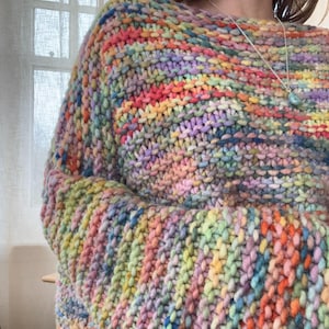Op de afbeelding: Een grof gebreide trui met een levendig, veelkleurig garenpatroon met gemengde tinten roze, geel, blauw, groen en oranje. Het kledingstuk heeft een brede, ontspannen halslijn en een dik, gestructureerd steekpatroon door de gehele stof heen.