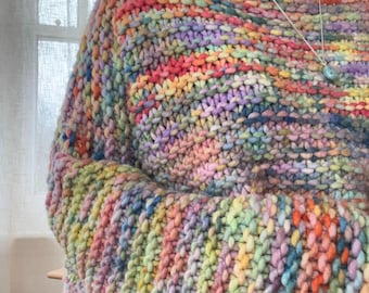 May include: A chunky knit sweater featuring a vibrant, multi-colored yarn pattern with blended hues of pink, yellow, blue, green, and orange. The garment has a wide, relaxed neckline and a thick, textured stitch pattern throughout the fabric.