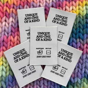 Op de afbeelding: Vijf witte stoffen labels met de tekst "UNIQUE AND ONE OF A KIND" in het zwart. De labels bevatten ook de zinnen "HAND WASH" en "DRY FLAT" met bijbehorende pictogrammen, en de tekst "JUST LIKE YOU!" op een kleurrijke gebreide achtergrond.