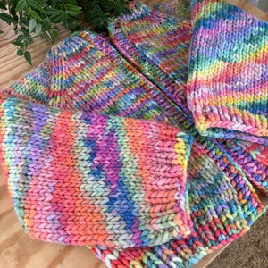 May include: A collection of hand-knitted items in vibrant rainbow colors. The items appear to be made of thick yarn and include what looks like a hat and a scarf. The knitted texture is clearly visible, with a mix of pink, orange, yellow, green, blue, and purple hues.