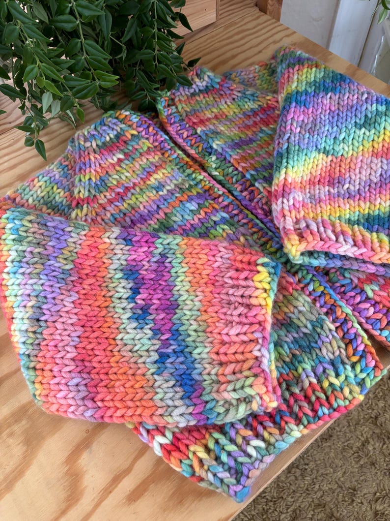 May include: A collection of hand-knitted items in vibrant rainbow colors. The items appear to be made of thick yarn and include what looks like a hat and a scarf. The knitted texture is clearly visible, with a mix of pink, orange, yellow, green, blue, and purple hues.