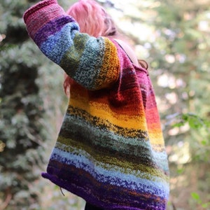 May include: A long-sleeved knit sweater featuring a horizontal gradient pattern of vibrant colors including purple, blue, green, yellow, orange, and red. The garment has a loose, relaxed fit with a wide neckline and rolled edges at the cuffs and hem.