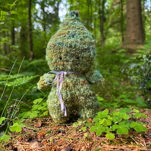 May include: A small, handmade crochet doll with a textured, variegated green and brown yarn body. The doll has a pointed head, two tiny black eyes, a simple stitched mouth, and a light purple ribbon tied around its neck with a small silver bell.