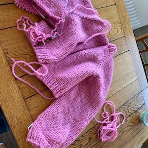 May include: A partially completed pink knitted garment, possibly a sweater, lies on a wooden table. Knitting needles are inserted into the work, with loose yarn and a knitted hat nearby. The project showcases a chunky knit stitch.