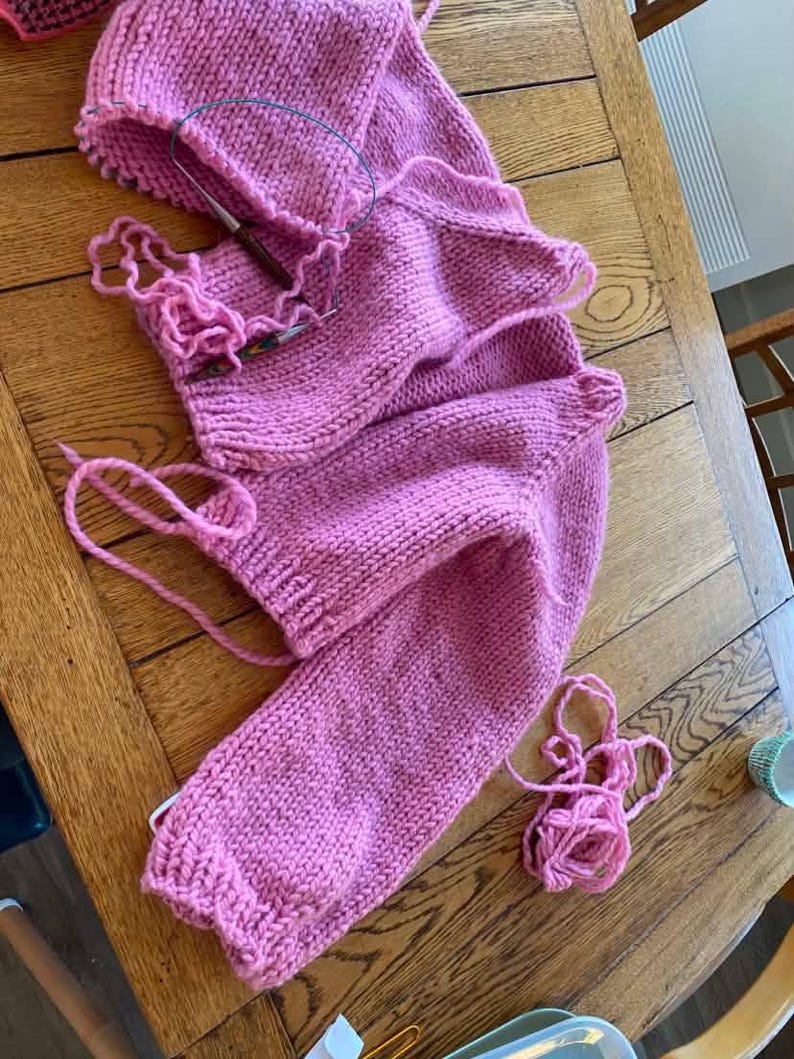 May include: A partially completed pink knitted garment, possibly a sweater, lies on a wooden table. Knitting needles are inserted into the work, with loose yarn and a knitted hat nearby. The project showcases a chunky knit stitch.