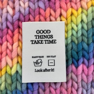 May include: Close-up of a vibrant, multicolored knitted fabric in shades of pink, purple, yellow, and green. A white label with the text "GOOD THINGS TAKE TIME" and care instructions is visible.