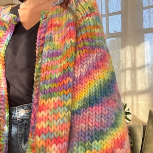 May include: A vibrant, rainbow-colored, chunky knit cardigan. The sweater features a spectrum of colors, including pink, yellow, green, blue, and purple. The thick yarn creates a textured, cozy appearance. The cardigan is worn over a black top and blue jeans.