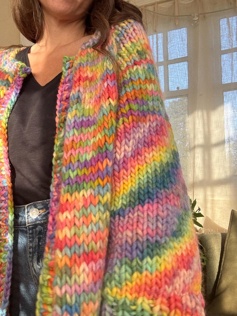 May include: A vibrant, rainbow-colored, chunky knit cardigan. The sweater features a spectrum of colors, including pink, yellow, green, blue, and purple. The thick yarn creates a textured, cozy appearance. The cardigan is worn over a black top and blue jeans.
