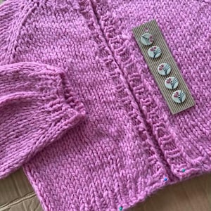 May include: A hand-knitted, light purple cardigan with a textured stitch pattern. The sweater features a button-up front and decorative buttons with floral designs. The sleeves and neckline have a detailed knit design.