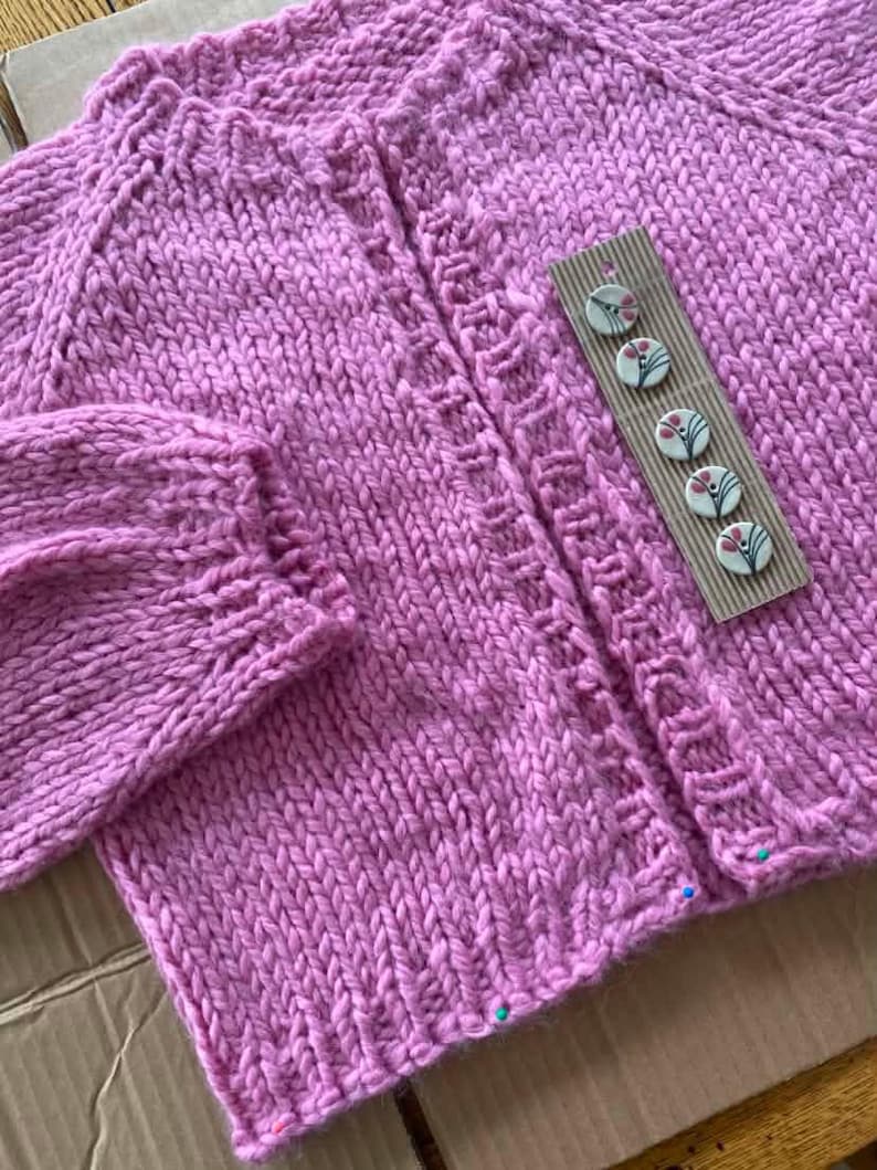 May include: A hand-knitted, light purple cardigan with a textured stitch pattern. The sweater features a button-up front and decorative buttons with floral designs. The sleeves and neckline have a detailed knit design.