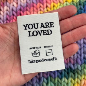 Könnte beinhalten: Ein weißes Stoffetikett mit dem schwarzen Schriftzug "YOU ARE LOVED". Es enthält Pflegehinweise: "HAND WASH DRY FLAT" und den Satz "Take good care of it". Das Etikett befindet sich auf einem bunten, regenbogenfarbenen Strickhintergrund.