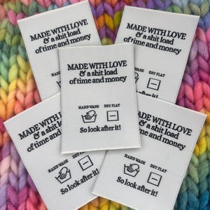 Può includere: Etichette in tessuto bianco con testo nero: "MADE WITH LOVE & a shit load of time and money." Ogni etichetta include anche simboli di lavaggio a mano e asciugatura in piano, e la frase "So look after it!" Le etichette sono su uno sfondo a maglia colorato.