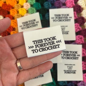 May include: Several white fabric labels with the text "THIS TOOK >>> FOREVER <<< TO CROCHET" in black. The labels are held over a colorful, crocheted blanket. Some labels also include a washing symbol and the word "FOREVER".