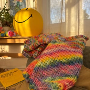 May include: A vibrant, hand-knitted rainbow-colored garment, possibly a sweater or blanket, is draped over a chair. The piece features a thick, textured knit with bands of pink, yellow, green, and blue. A yellow smiley face ball and a book titled "Butterflies" are in the background.