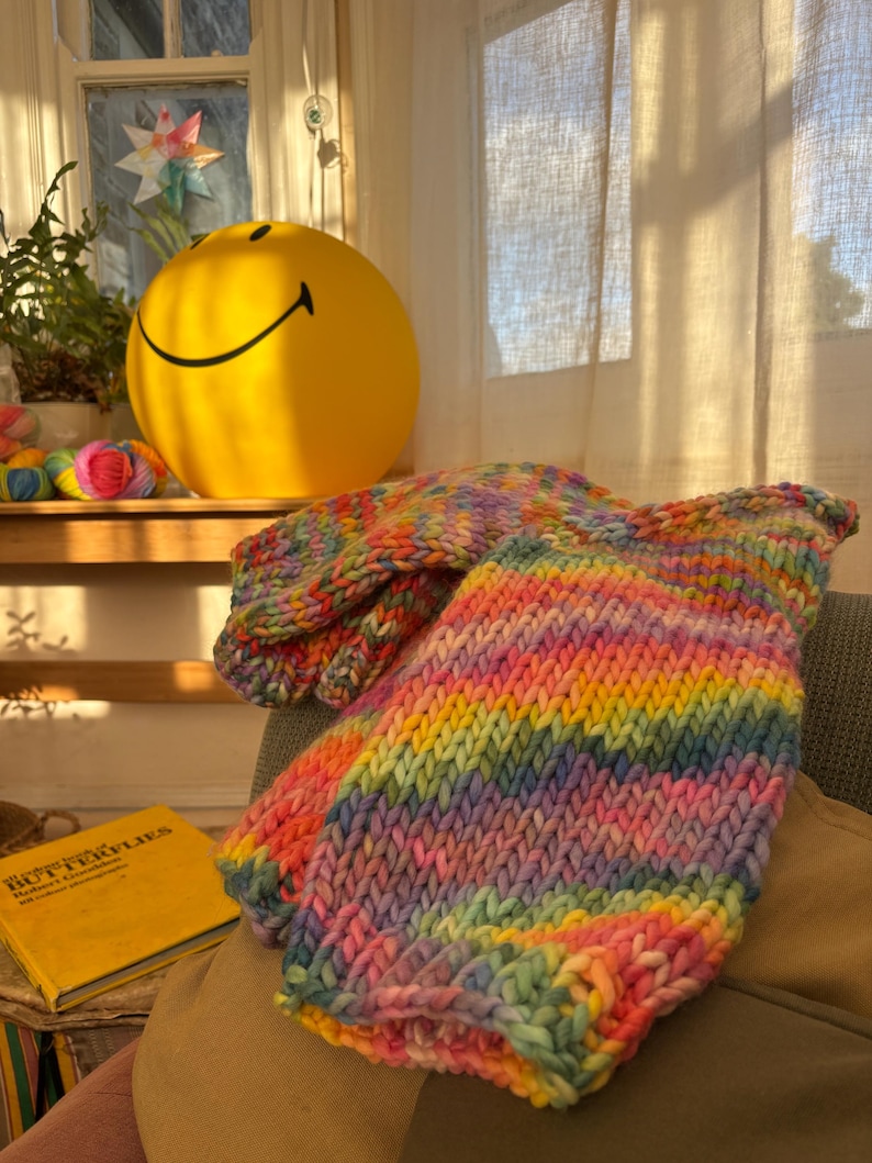 May include: A vibrant, hand-knitted rainbow-colored garment, possibly a sweater or blanket, is draped over a chair. The piece features a thick, textured knit with bands of pink, yellow, green, and blue. A yellow smiley face ball and a book titled "Butterflies" are in the background.