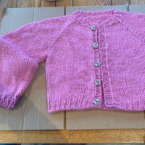 May include: A hand-knitted, light pink cardigan with a round neckline and long sleeves. The sweater features wooden buttons and a ribbed hem and cuffs. The garment is displayed on a cardboard surface.