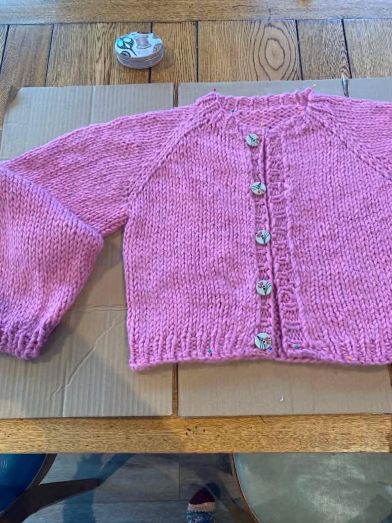 May include: A hand-knitted, light pink cardigan with a round neckline and long sleeves. The sweater features wooden buttons and a ribbed hem and cuffs. The garment is displayed on a cardboard surface.