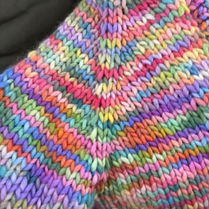 May include: Close-up of a hand-knitted garment in a vibrant array of colors. The chunky knit features a mix of pinks, purples, oranges, yellows, and greens. The texture of the yarn is clearly visible, creating a cozy and textured appearance.