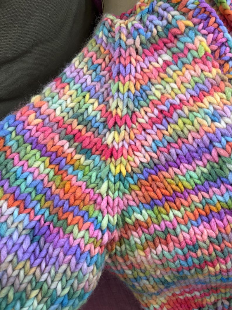 May include: Close-up of a hand-knitted garment in a vibrant array of colors. The chunky knit features a mix of pinks, purples, oranges, yellows, and greens. The texture of the yarn is clearly visible, creating a cozy and textured appearance.