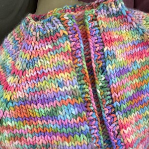 May include: A chunky knit cardigan in a vibrant rainbow of colors, including pink, yellow, green, and blue. The thick yarn creates a textured look, with a visible stitch pattern. The open front design and high collar add to the cozy aesthetic.