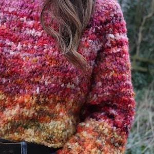 May include: A textured, colorful sweater in shades of pink, red, orange, and yellow. The sweater has a crew neck and long sleeves. The sleeves and lower portion of the sweater have a gradient effect, transitioning from yellow to orange.