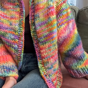 Może przedstawiać: Gruby, tęczowy kardigan z dzianiny. Sweter z otwartym przodem ma żywą mieszankę kolorów, w tym różowy, pomarańczowy, żółty, zielony i niebieski. Gruba przędza tworzy teksturowany, przytulny wygląd, idealny do dodania koloru do każdego stroju.